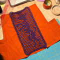 An orange garment laid flat on a wooden desk, with a purple crocheted lace panel attached along the front. Surrounding the garment are sewing tools and supplies, including a ruler, tape, pins, and a laptop.