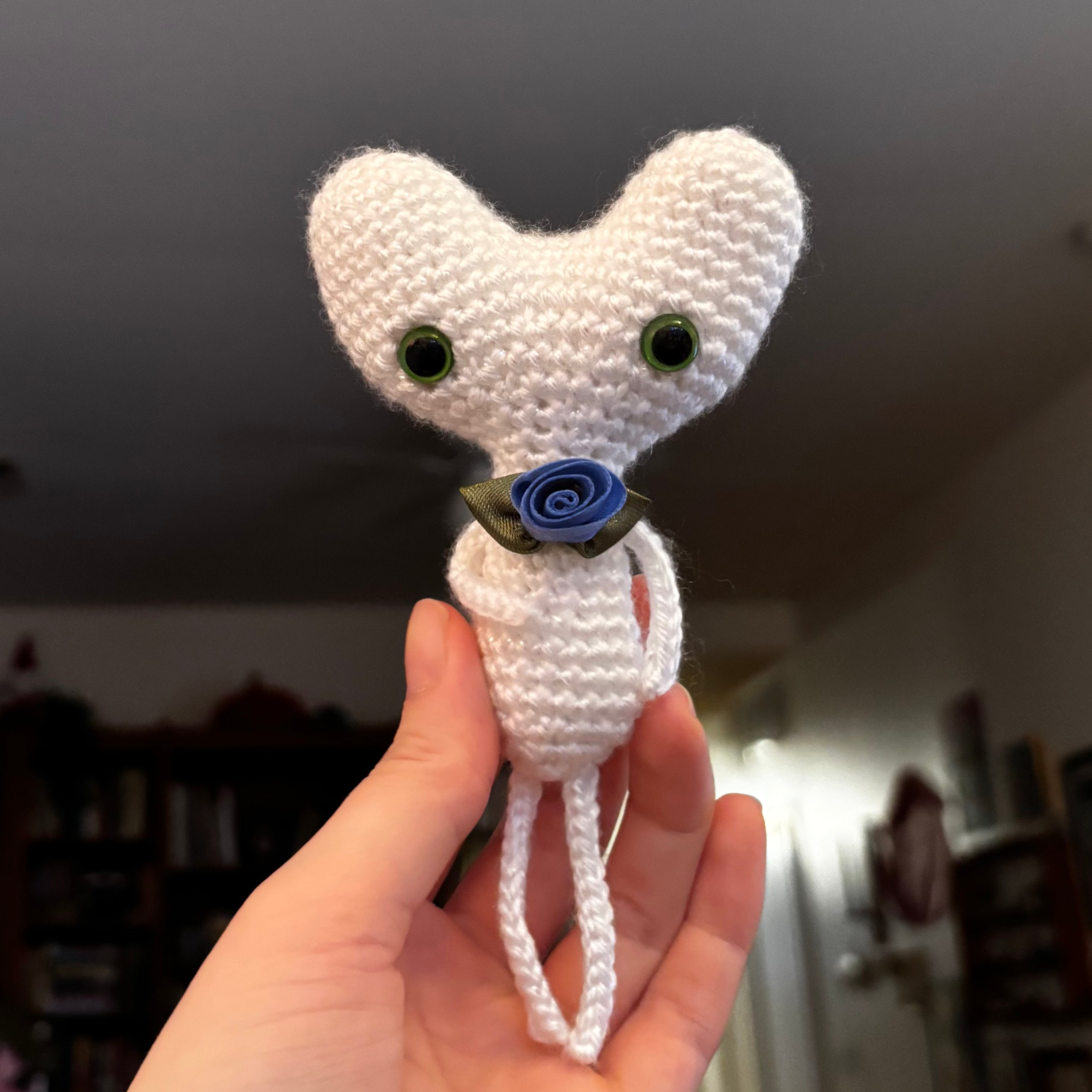 Hand holding a small white crocheted Cry Buddy toy with green eyes and a blue flower on a neutral background.